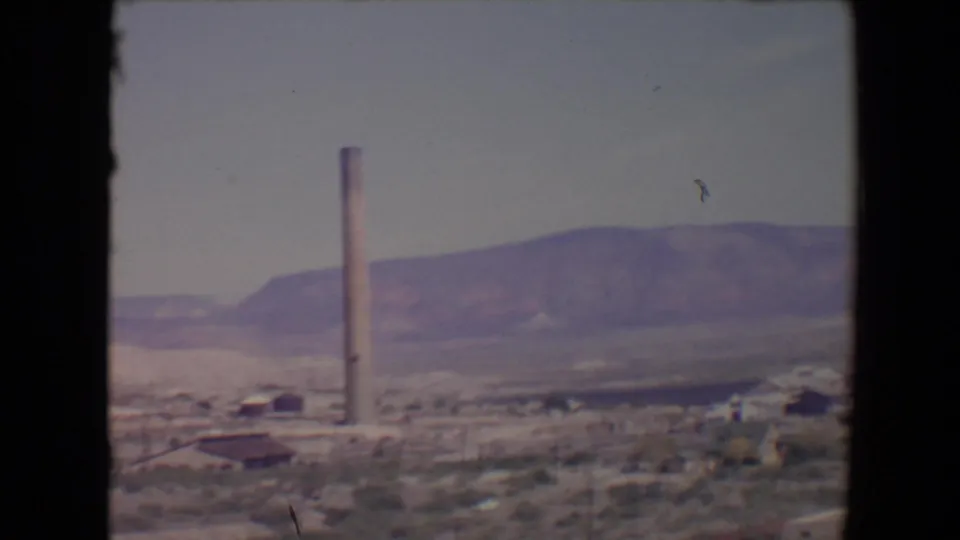 8mm home-movie frame of Flagstaff, Arizona, showing a smokestack, low structures, sparse vegetation, and a broad mountain ridge.