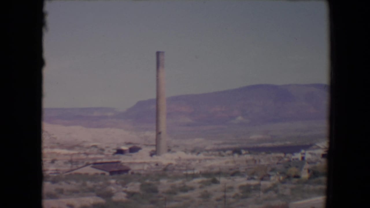1967 8mm view near Flagstaff, Arizona, showing a tall smokestack, low buildings, scrubland, and distant mountains.
