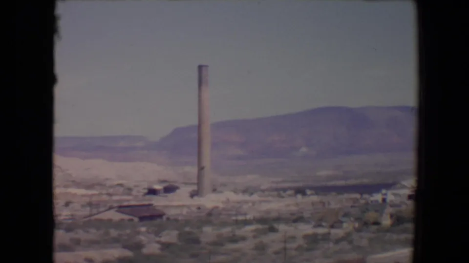1967 8mm view near Flagstaff, Arizona, showing a tall smokestack, low buildings, scrubland, and distant mountains.