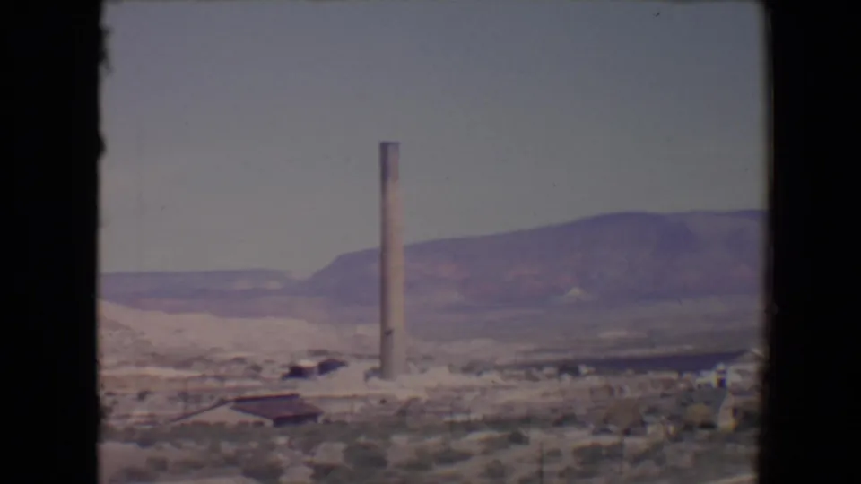 Flagstaff-area 1967 home-movie frame with a vertical smokestack, small buildings, dry terrain, and mountain backdrop.