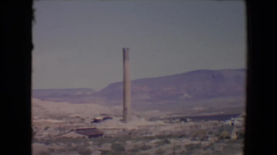 1967 8mm still of a tall industrial smokestack in a desert setting near Flagstaff, Arizona, with mountains behind it.