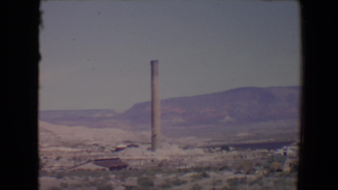 1967 Flagstaff, Arizona 8mm frame showing a tall smokestack, sparse desert foreground, low structures, and hills under a pale sky.