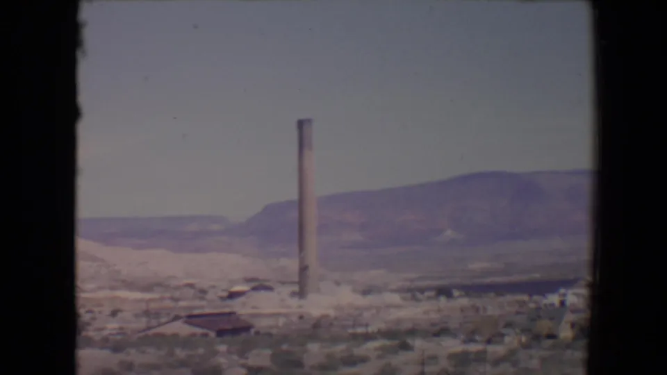 1967 Flagstaff, Arizona 8mm frame showing a tall smokestack, sparse desert foreground, low structures, and hills under a pale sky.