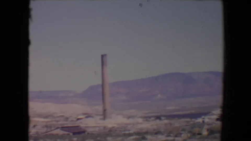 Flagstaff, Arizona home-movie frame with a tall smokestack centered in an open desert landscape and muted hills in the background.