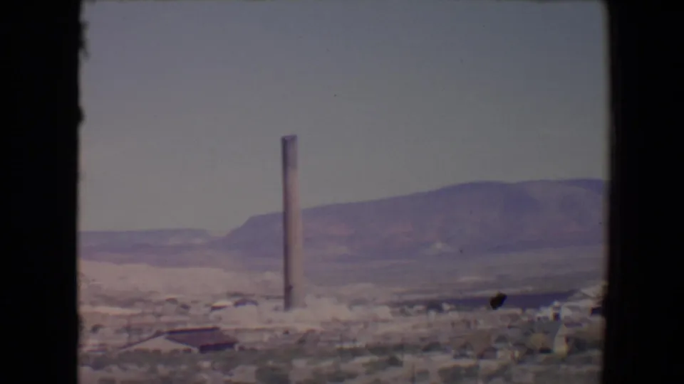 1967 8mm view of a smokestack and small structures in a Flagstaff desert setting, framed by distant hills and a pale sky.
