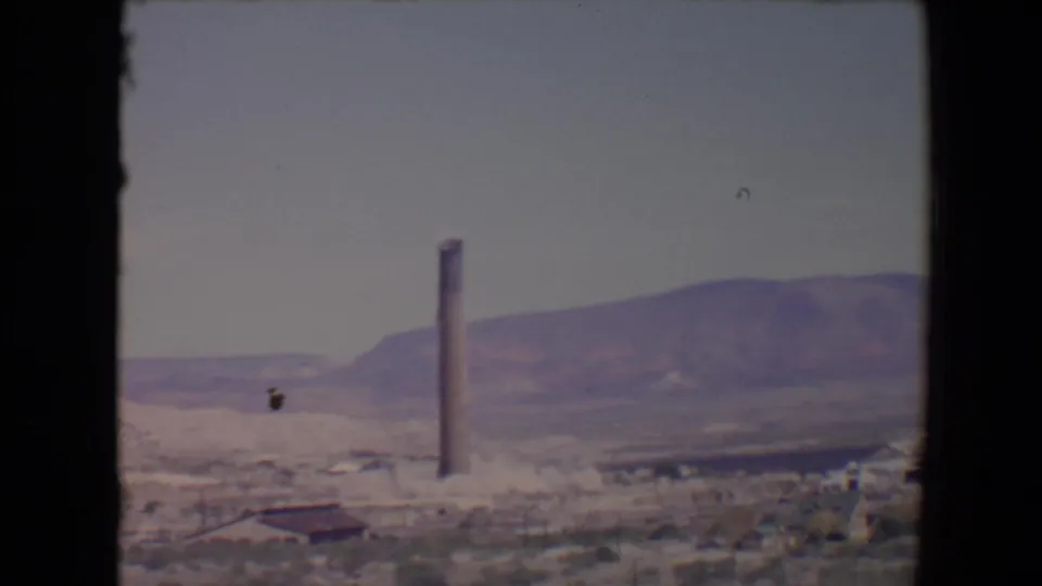 Flagstaff, Arizona 8mm frame showing the smokestack slightly right of center, with open terrain, low structures, and distant hills.