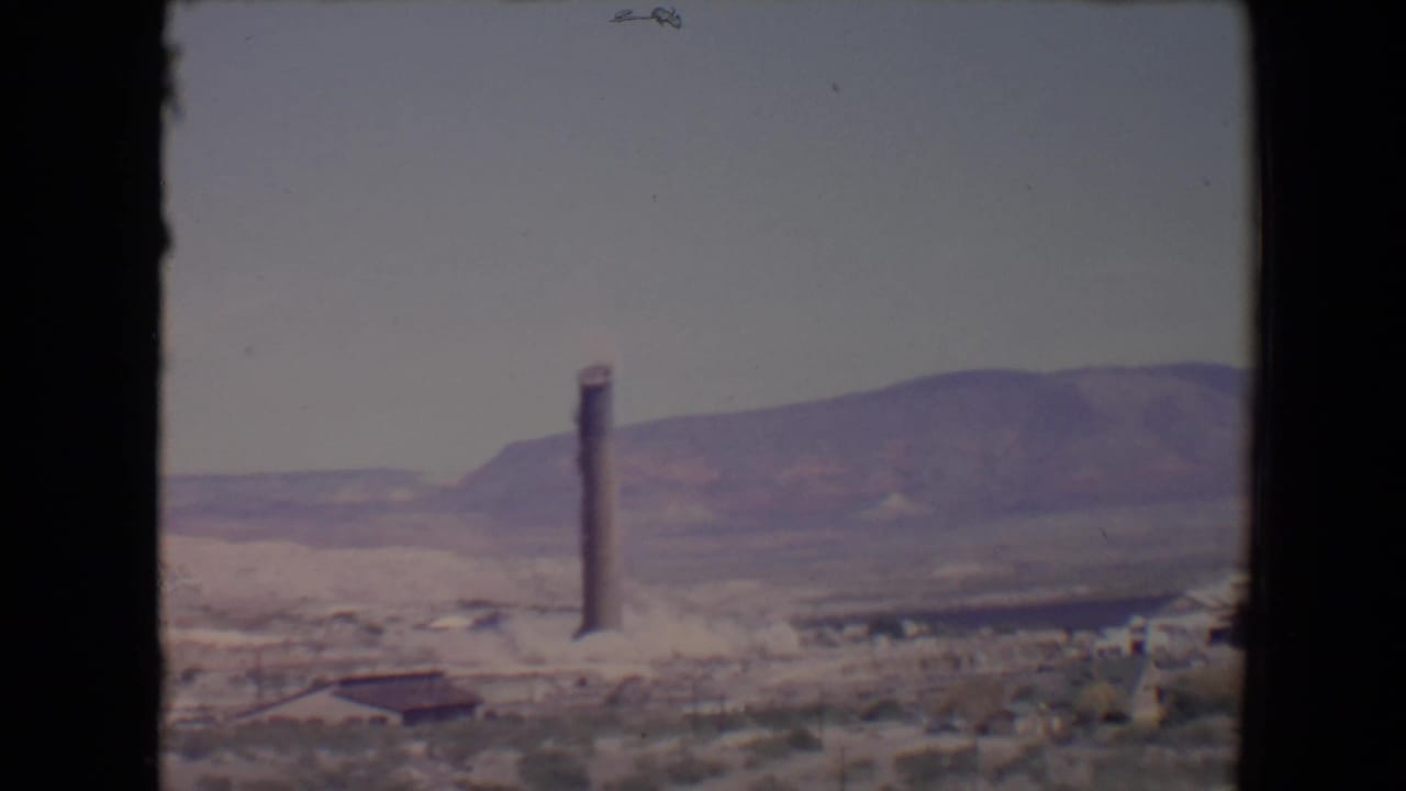 1967 Flagstaff-area 8mm frame showing a tall smokestack or tower before collapse, set against distant mountains and scrubby ground.