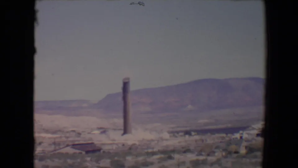 1967 Flagstaff-area 8mm frame showing a tall smokestack or tower before collapse, set against distant mountains and scrubby ground.