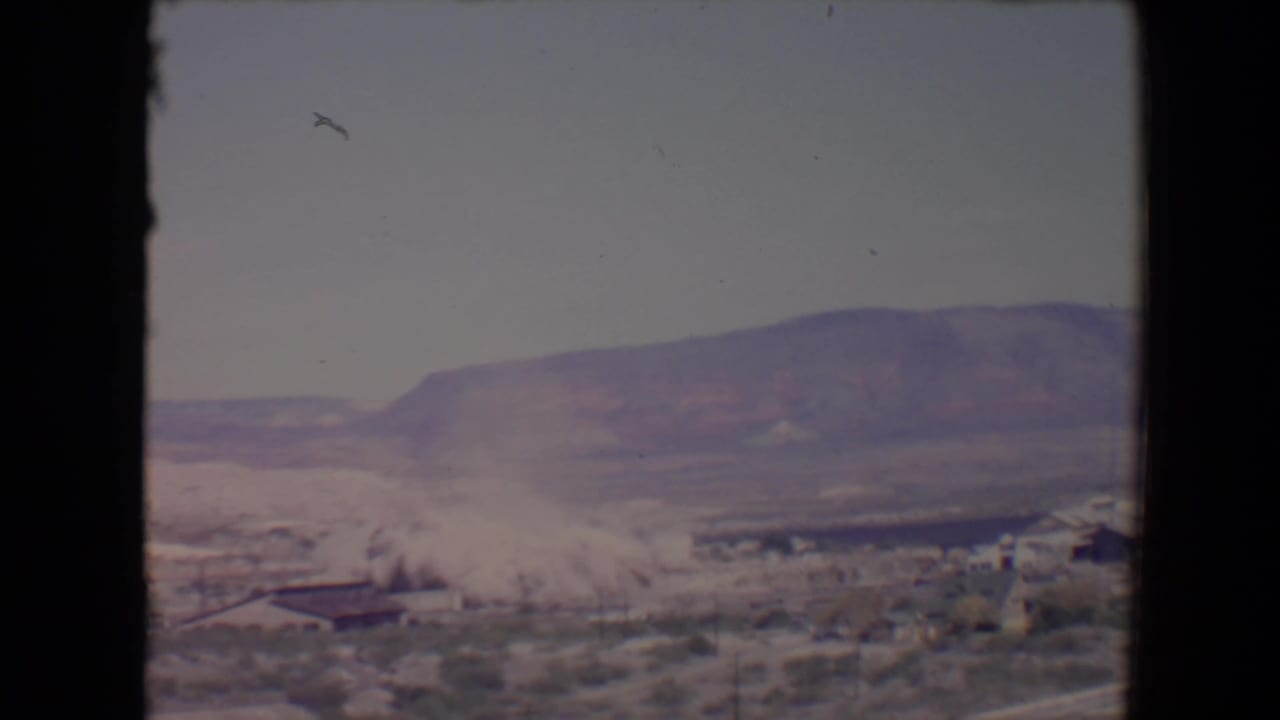 Dust cloud beginning to rise near low buildings with mountains in the distance.
