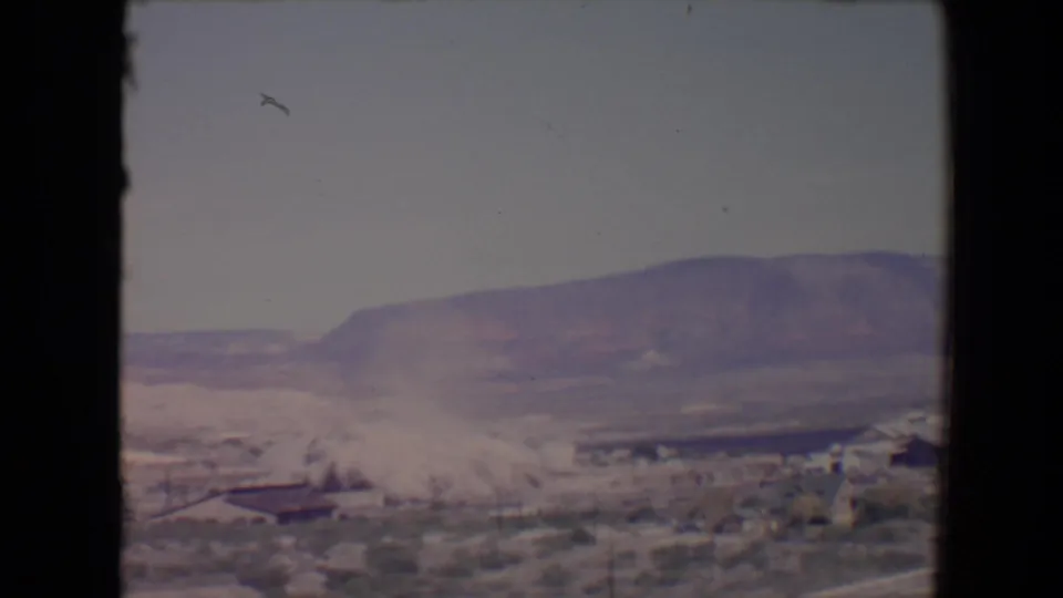 1967 Flagstaff-area 8mm view of a demolition dust cloud starting to spread across low desert-town structures below distant mountains.