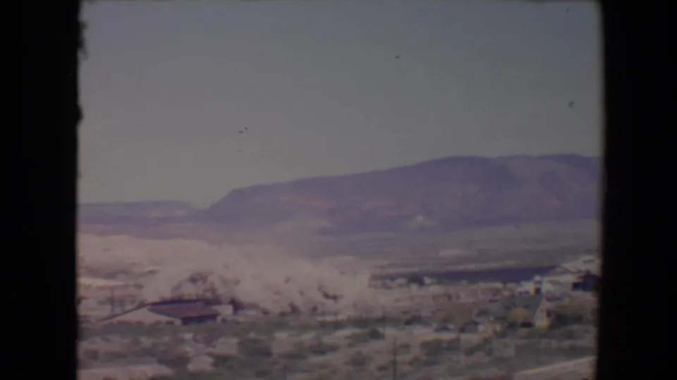 A thick demolition dust cloud hangs over low buildings and scrubland in the 1967 Flagstaff footage, with a broad mountain ridge behind.