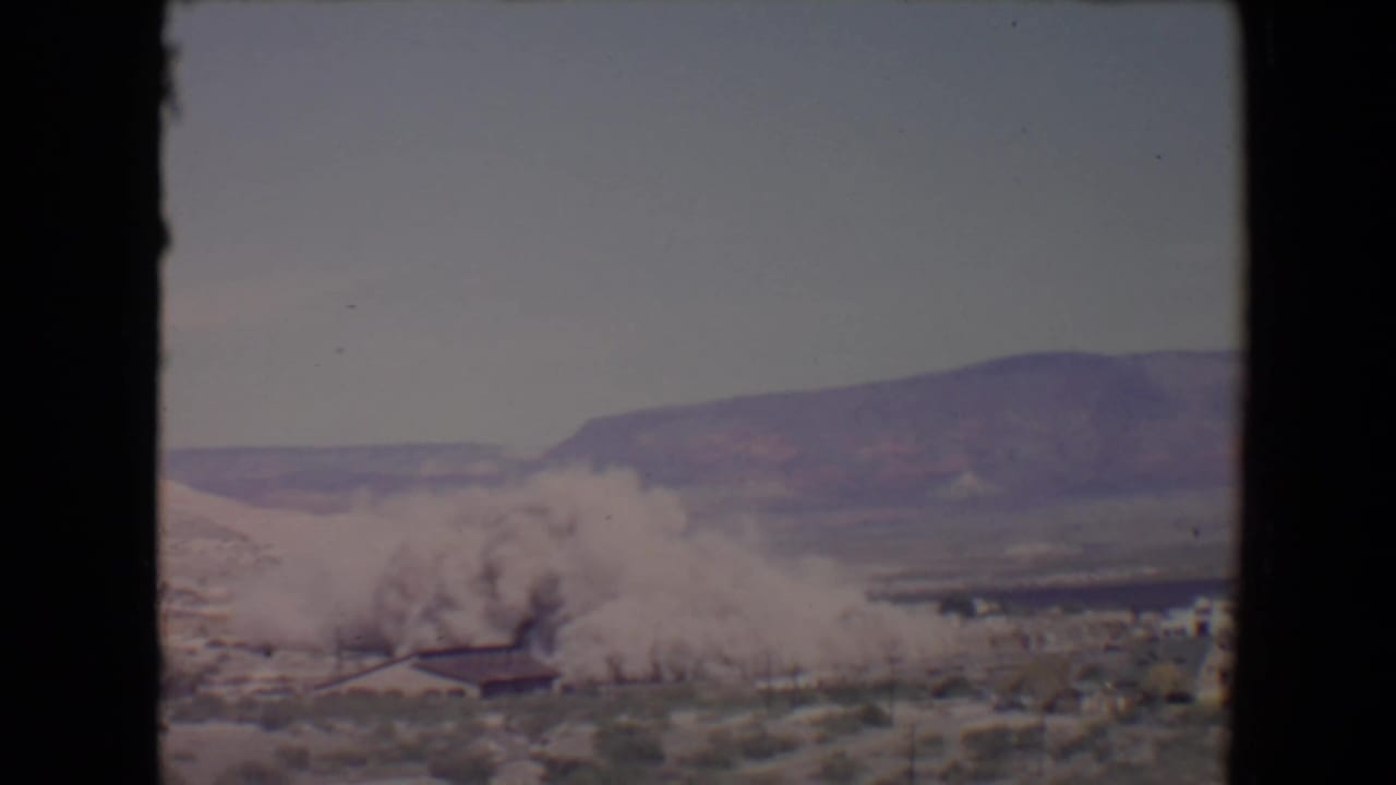 Distant smoke plume rising near a low building in a desert landscape.