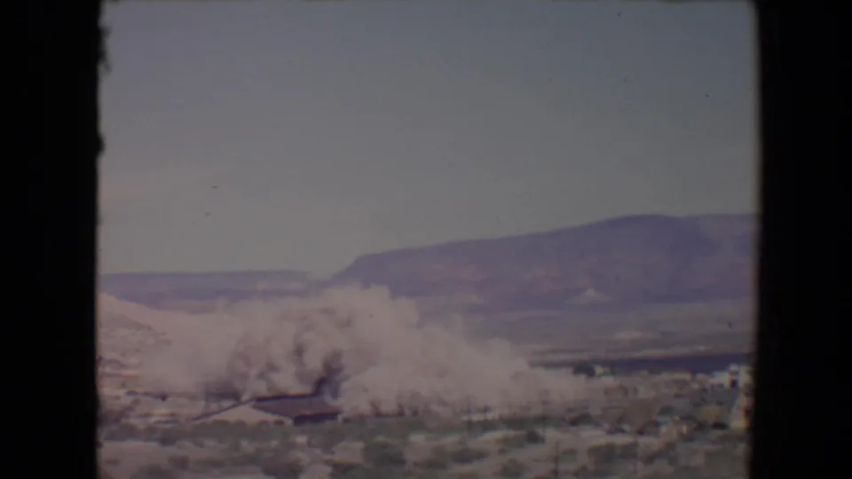 1967 Flagstaff, Arizona 8mm frame showing smoke or dust beginning to spread near a low structure with hills behind it.