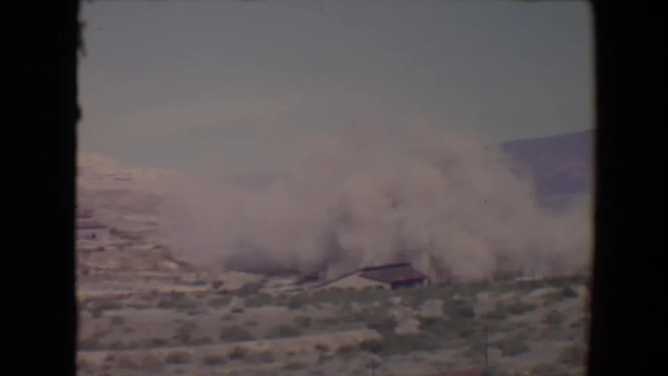 1967 Arizona 8mm frame with a broad smoke plume drifting across desert ground near a small building, seen through dark film borders.