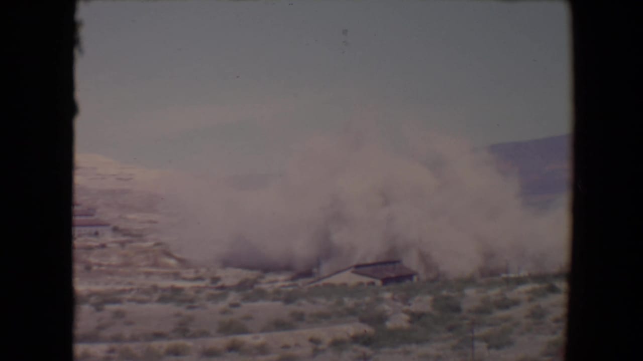 Large smoke plume rising near low buildings in a desert landscape with hills beyond.