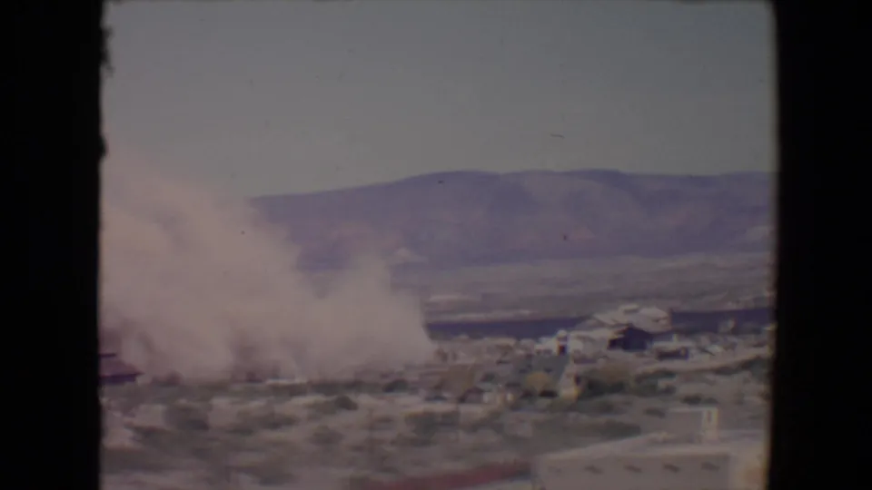1967 Flagstaff, Arizona frame with smoke drifting across low buildings, desert ground, and a distant hillside.