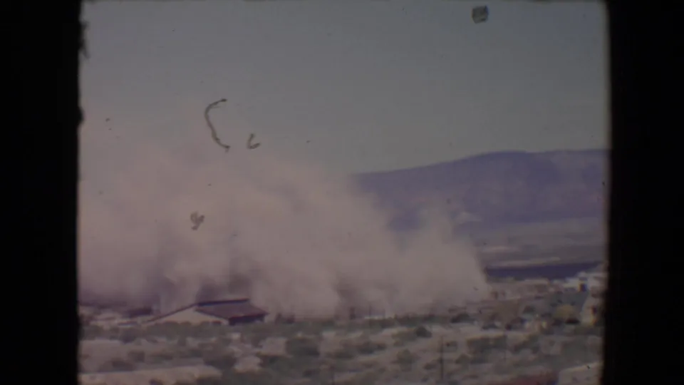 1967 Flagstaff-area 8mm frame showing a dust plume beginning to spread near a low desert structure.