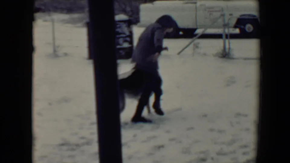 In a 1967 Flagstaff home-movie frame, a child in winter clothing bends toward a dog beside snow, porch framing, and a white pickup.