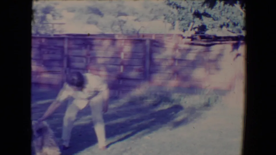 The man bends close to the lawn while the dog appears near the lower edge, captured with faded 8mm color and glare.