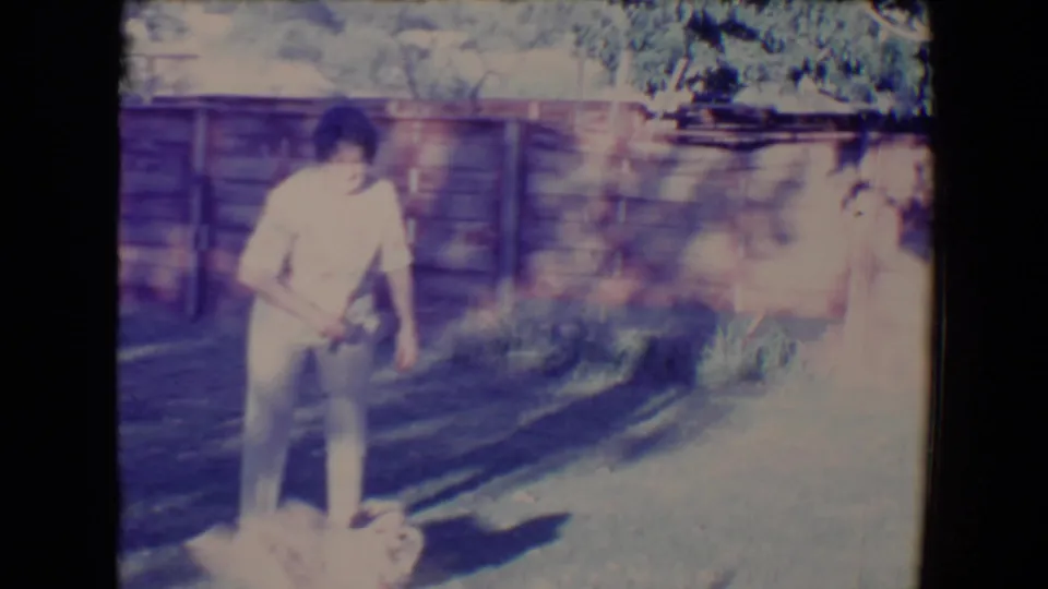 The man stands over the small fluffy dog in the Tucson backyard, with long shadows and a wooden fence in view.