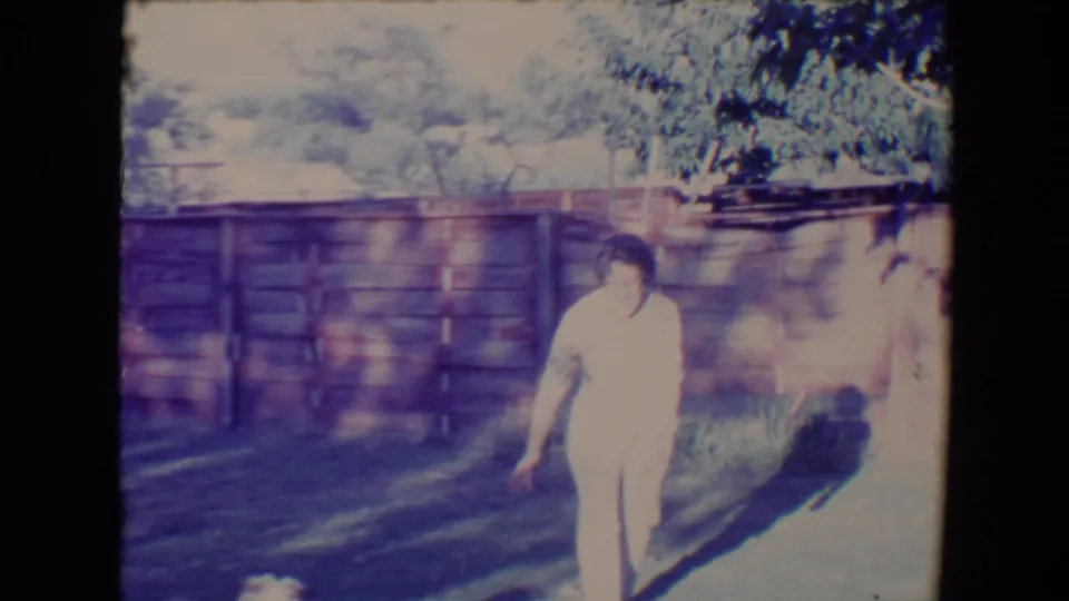 The woman walks through the sunlit Tucson backyard as the small dog remains low in the frame near the grass.