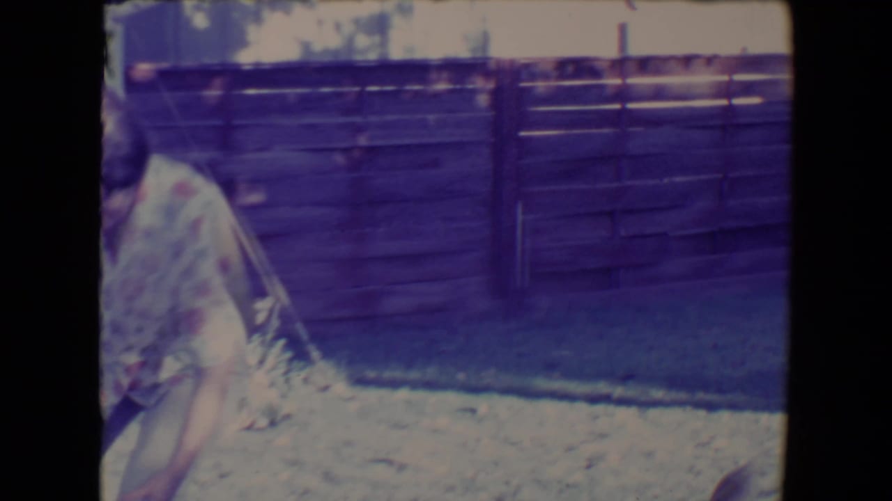 In the supplied 1967 Tucson home-movie context, a woman stands partly cropped beside a pale ball in a fenced backyard.