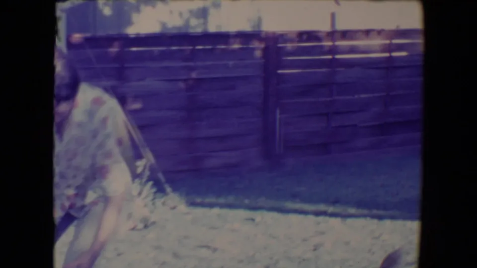 In the supplied 1967 Tucson home-movie context, a woman stands partly cropped beside a pale ball in a fenced backyard.