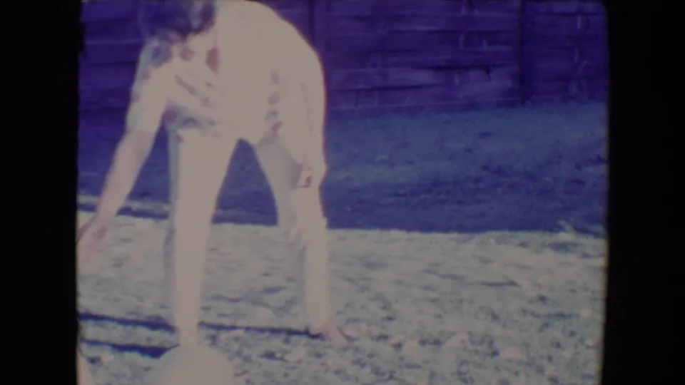 Woman bends down toward the pale ball on the lawn, continuing the backyard play in the 1967 Tucson home-movie footage.