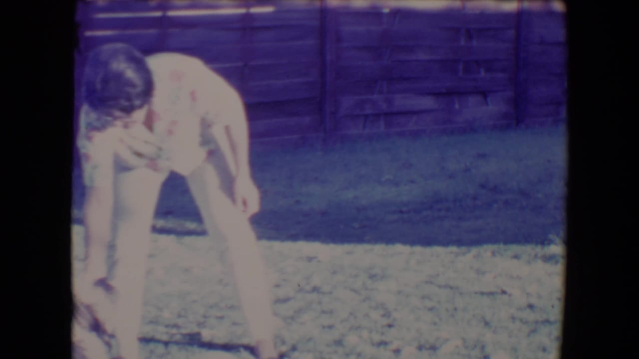 Woman bending toward a small dog on grass beside a wooden fence.