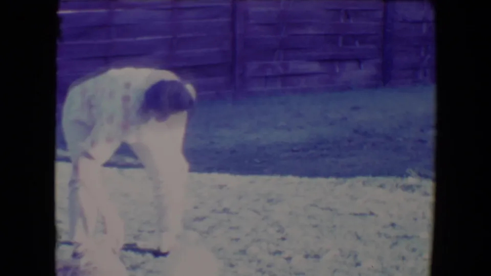 The woman remains bent over in the Tucson yard, with the small dog low in frame against grass and a wooden fence background.