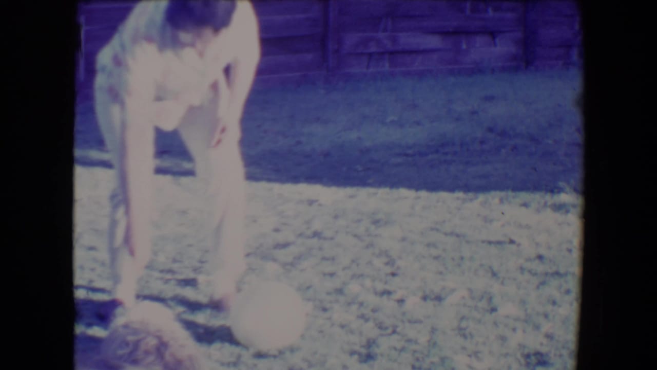 In a 1967 Tucson home-movie frame, an older woman in light clothing bends near a ball while a small dog stands on the lawn.