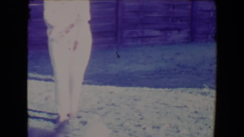 The woman stands in the Tucson backyard with the ball near her feet, framed by grass, fence boards, and aged 8mm color shift.