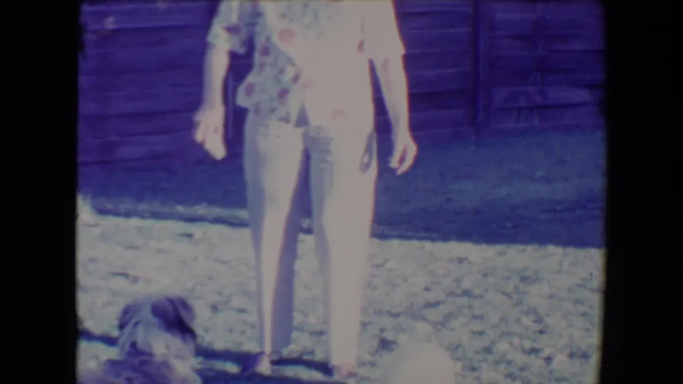 A small dark dog approaches the woman’s feet in the 1967 Tucson backyard scene, with a pale ball nearby.