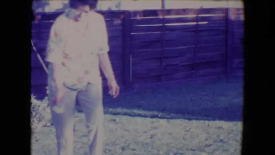 Adult woman in a floral shirt and light pants stands in a Tucson backyard on 1967 8mm home-movie footage.