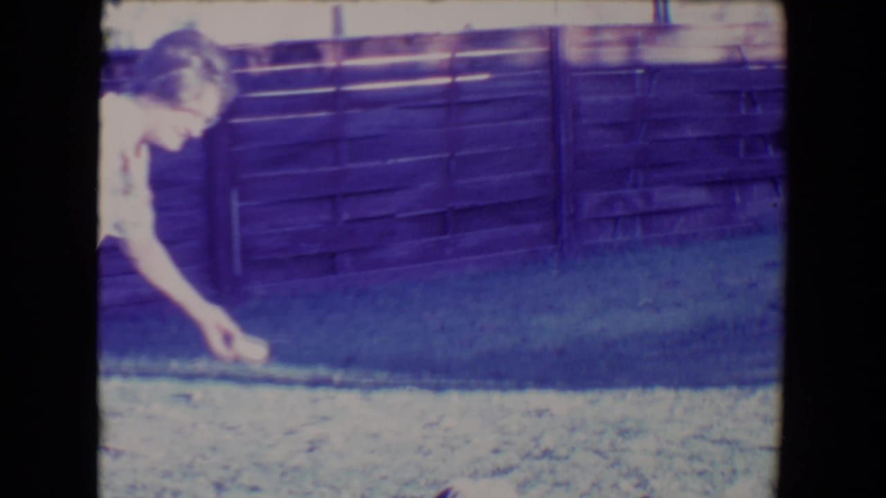 Adult woman bends over the lawn in a backyard, likely picking up or tossing a small fetch object.