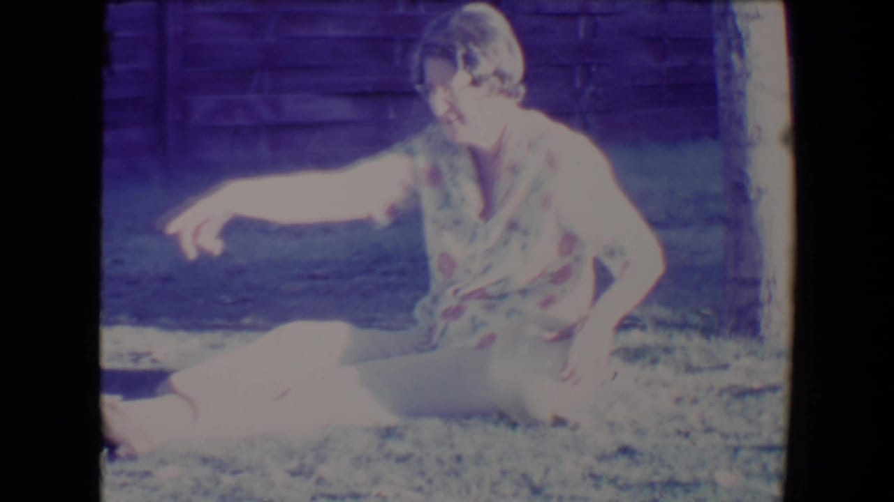 Woman seated on grass reaching toward a light-colored dog near a tree and wooden fence.