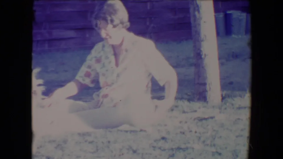 A woman in a floral dress sits cross-legged on a Tucson lawn, looking toward her dog near the edge of the frame.