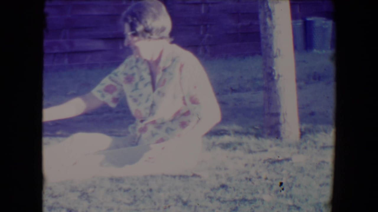 Elderly woman in a floral dress sits on grass beside a wood wall in bright sunlight.
