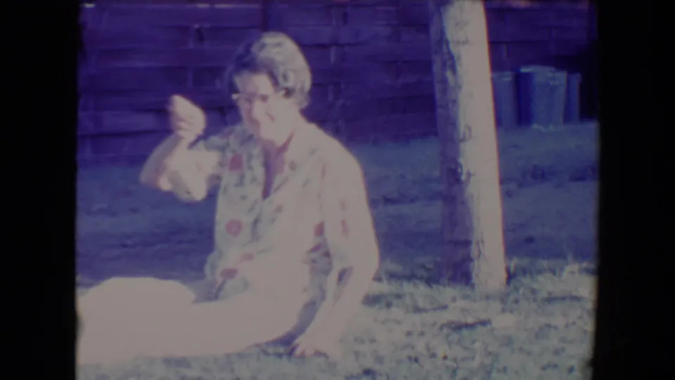 In the 1967 Tucson backyard scene, the woman lifts one hand as if throwing or showing an object while seated on the lawn.