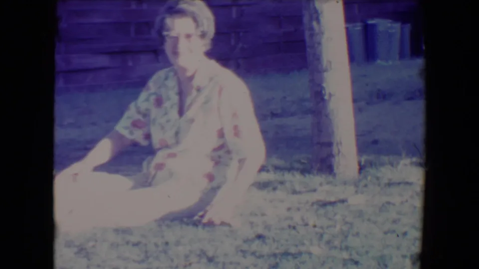 The 1967 Tucson home-movie frame shows the woman seated on the backyard grass, facing the camera in bright, overexposed sunlight.