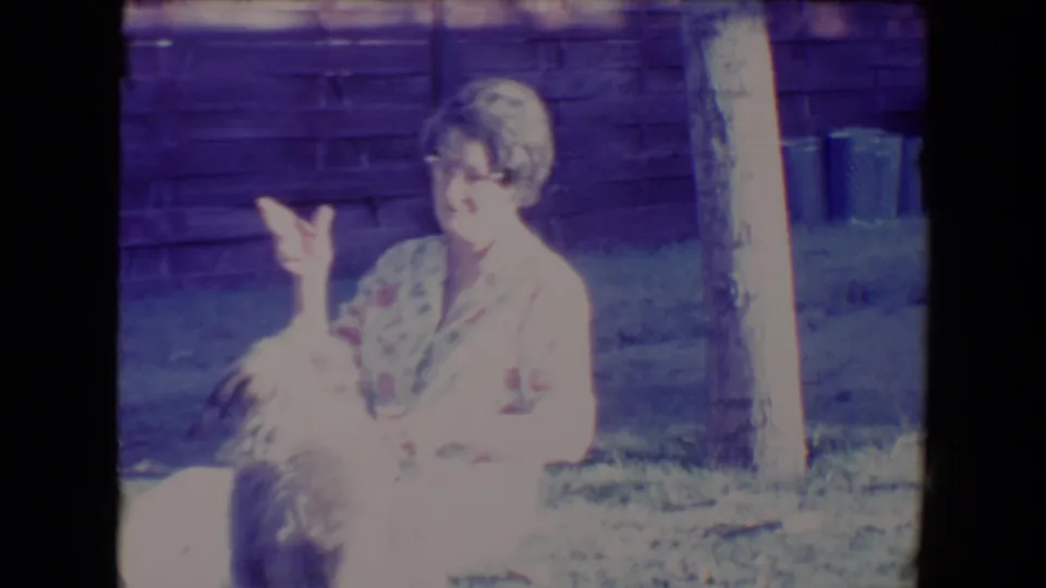 A woman in a floral dress raises her hand while seated beside a fluffy dog in the 1967 Tucson backyard scene.