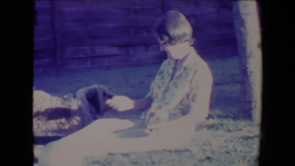The woman sits sideways on the lawn facing the dog, with a wooden fence and tree trunk visible in the Tucson home-movie frame.