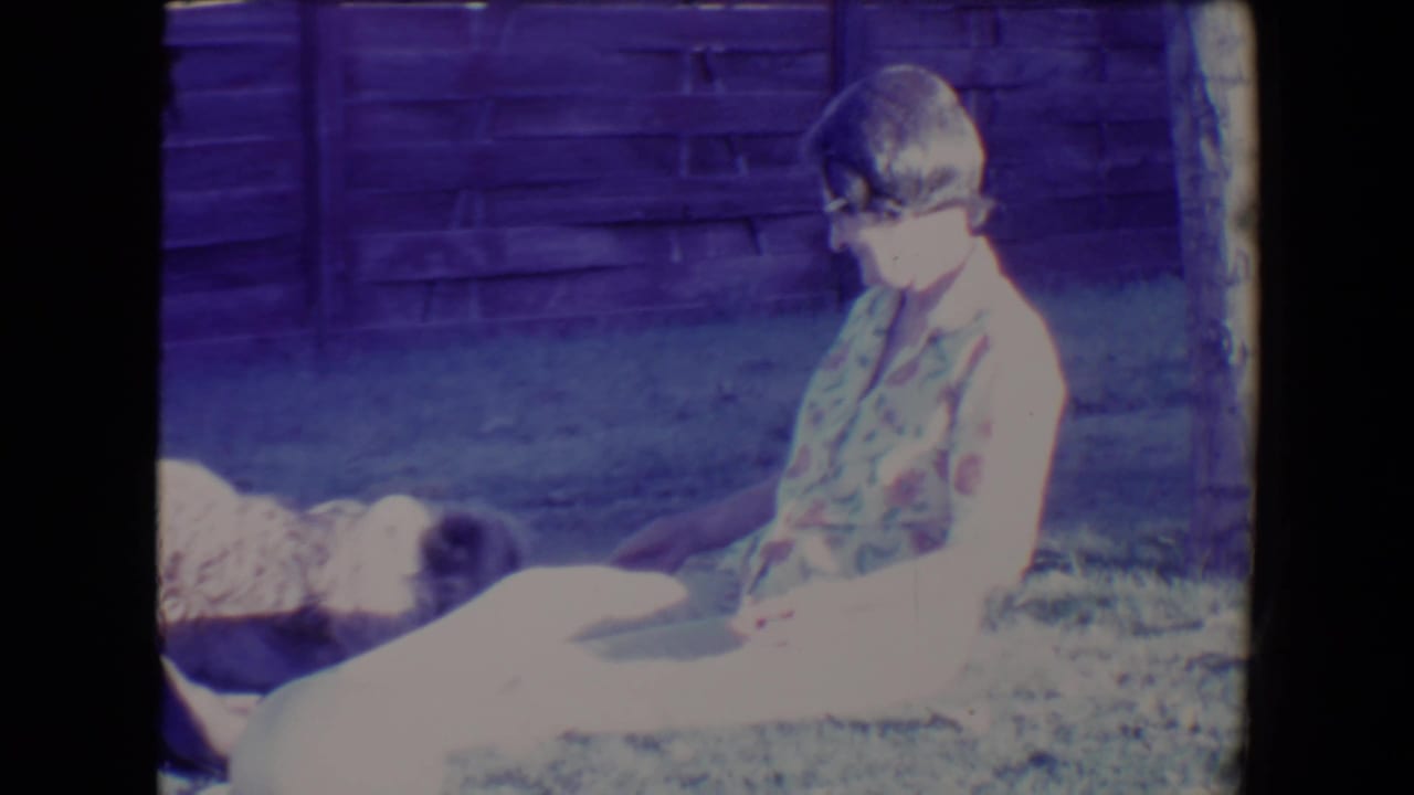 Woman seated on grass beside a small dog near a wooden fence.