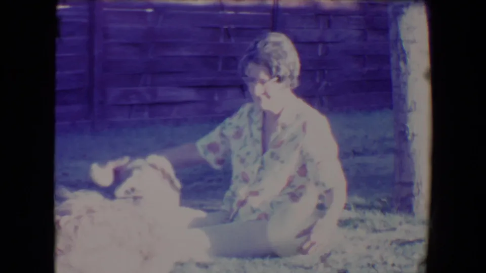 The woman reaches toward the dog beside her on the Tucson lawn, with a fence behind them and a soft purple 8mm cast.