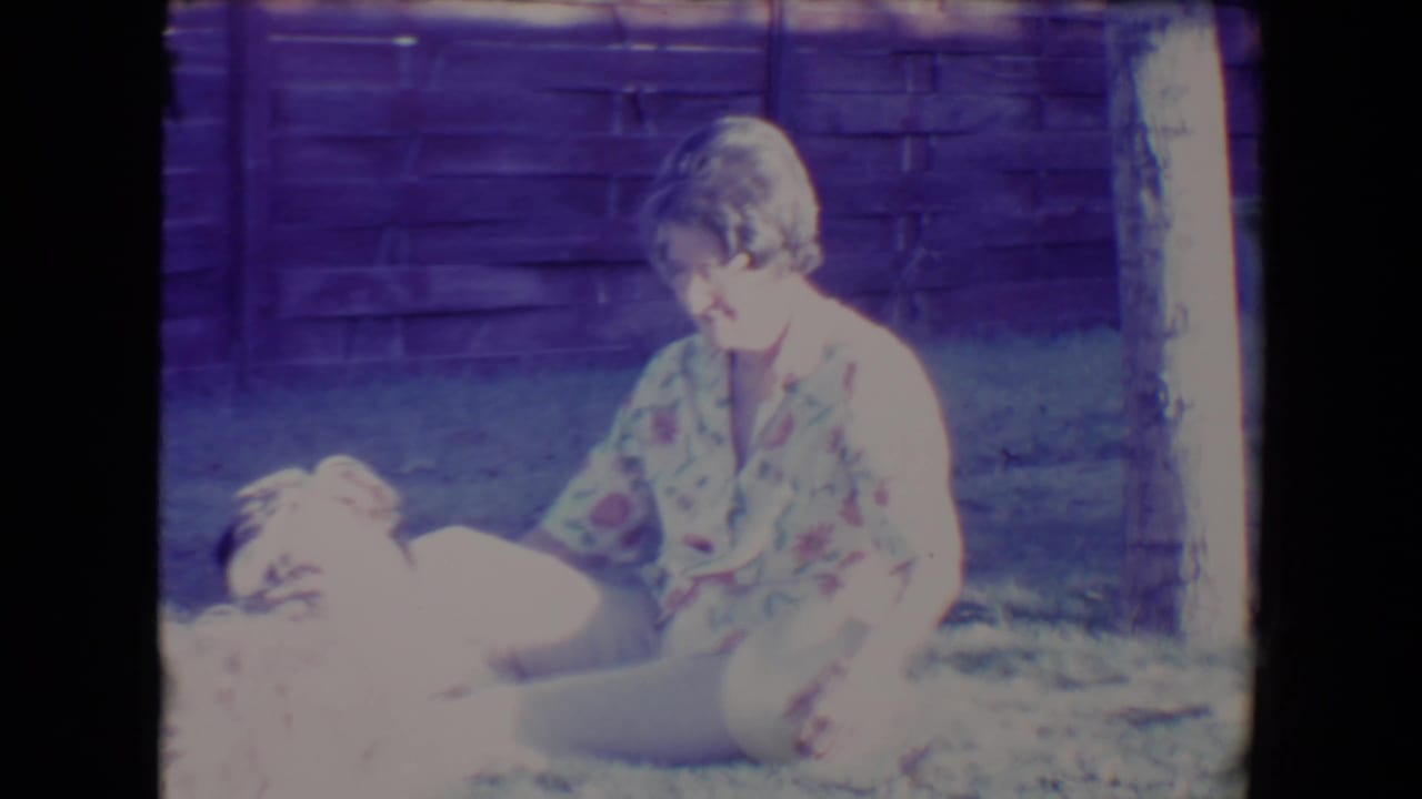 Woman sitting on grass beside a small fluffy dog in a fenced backyard.