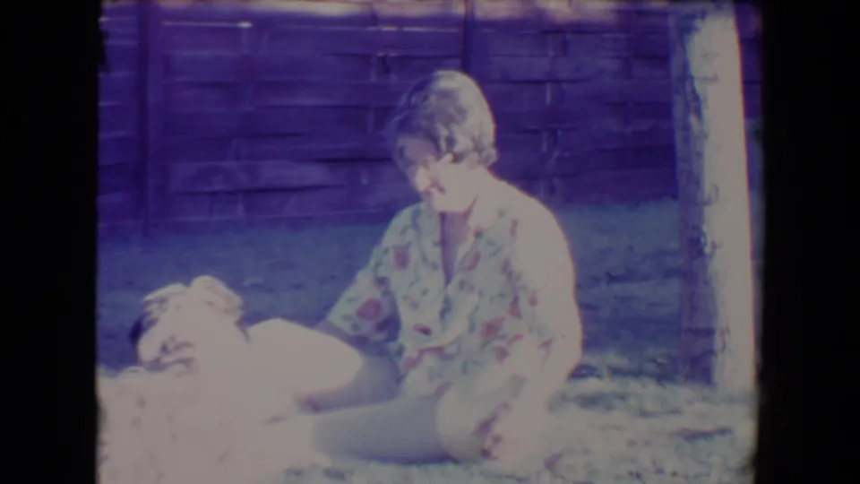 1967 Tucson home-movie frame showing a woman on a lawn with a small fluffy dog beside her near a wooden fence.