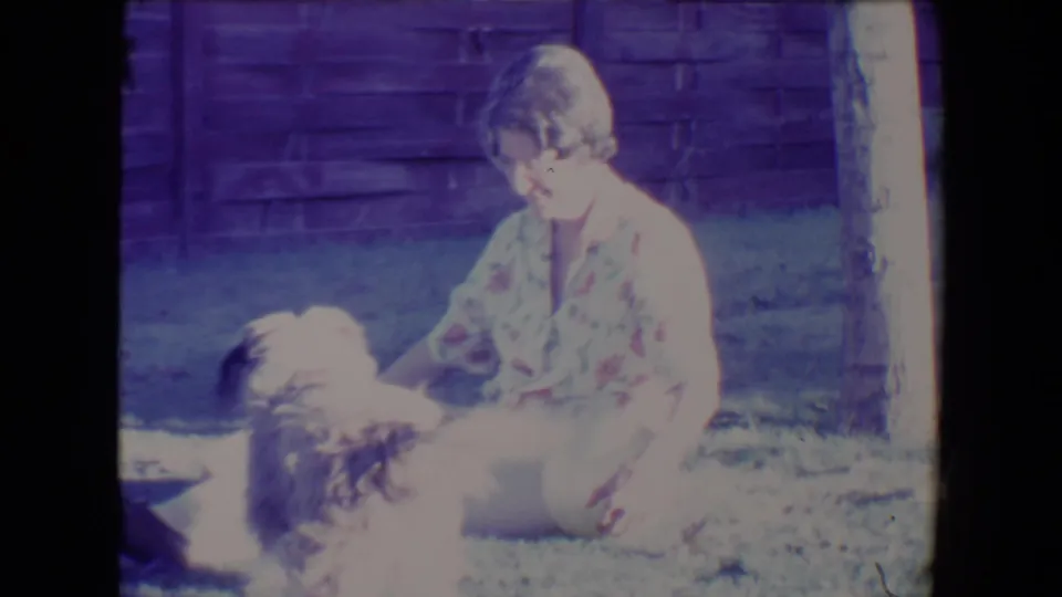 1967 Tucson backyard frame with a woman seated on grass as a small fluffy dog rolls or moves close to her.