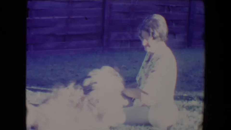 1967 Tucson home-movie frame showing the woman in profile as the small dog blurs with motion on the lawn.