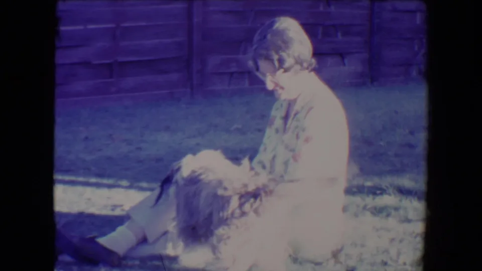 1967 Tucson 8mm frame showing a woman seated in a sunny yard with a fluffy dog close to her lap by a wooden fence.