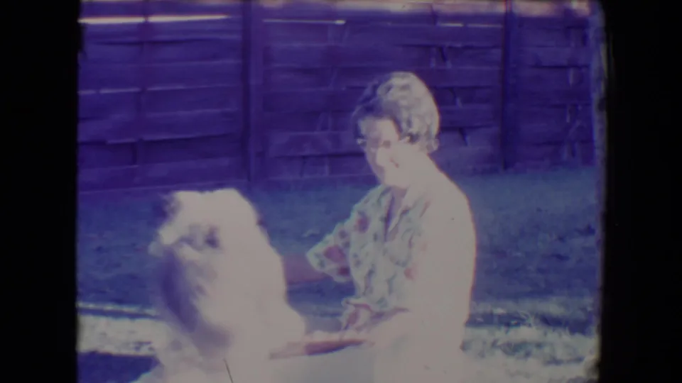 1967 Tucson home-movie frame with a fluffy dog sitting upright beside a seated woman in a sunny grassy yard.
